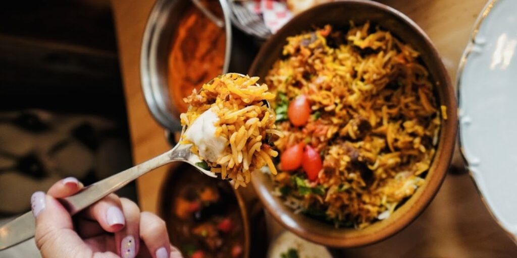 A person holding a spoon over a bowl of aromatic biryani, with saffron, cardamom, and cloves adding a unique fragrance and taste.