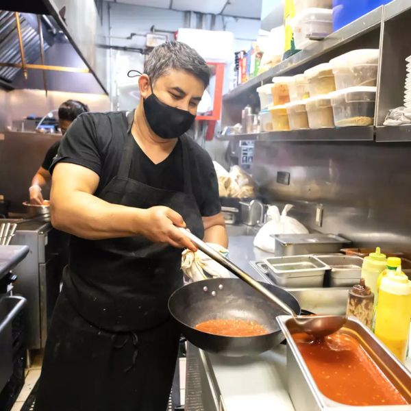 A man in a face mask and black apron skillfully prepares traditional Indian dishes in a vibrant kitchen setting.